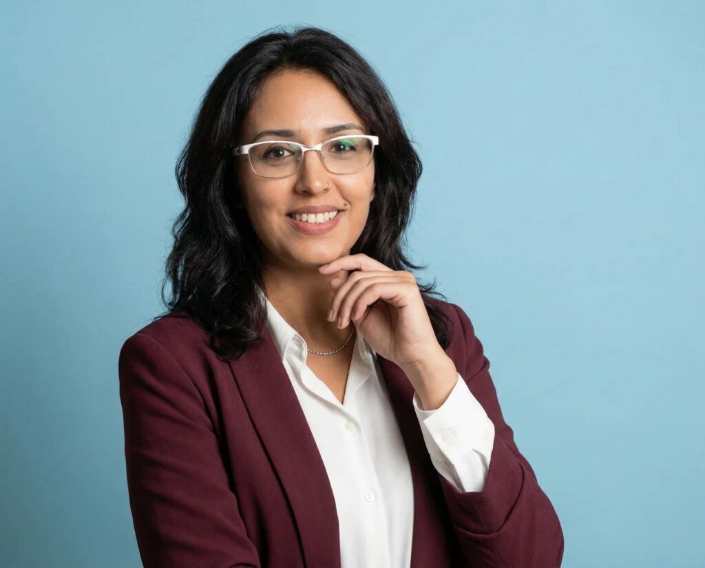 Ikram Foto mit weißer Bluse und Rotwein Jake, steht vor einer hellen blauen Hintergrund